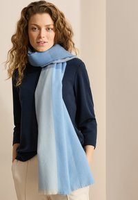 Scarf azzurro chiaro realizzato in un tessuto morbido, caratterizzato da un design a gradiente con estremità più chiare e bordi frangiati, indossato sopra un top scuro.