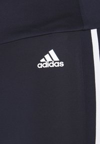 Mörkblå träningsleggings tillverkade av stretchigt tyg, med en vit Adidas-logotyp och vita sidostreifen längs de yttre sömmarna.