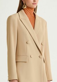 Blazer doppiopetto beige con due tasche frontali, realizzato in tessuto liscio. Abbinato a un dolcevita arancione a pois a contrasto.