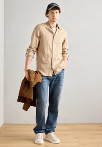 Cămașă bej din in cu butoni, tricou gri deschis, blugi albaștri, jachetă din suede maro, adidași albi, pălărie bleumarin cu text, stând pe un podea din lemn.