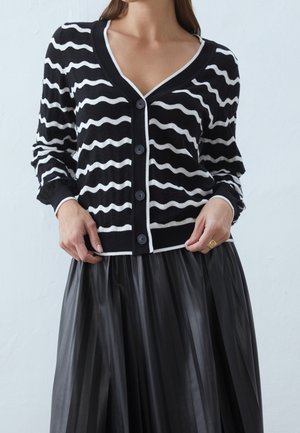 Vrouw die een zwart-wit golvend gestreepte cardigan met knopen draagt en een zwarte geplooide rok, terwijl ze de zoom van de cardigan met haar handen aanpast.