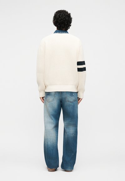 Personne de dos portant un pull en maille blanc avec des rayures noires sur la manche gauche et un jean bleu délavé et ample.