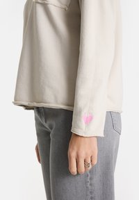 Person in einem hellbeigen Langarmshirt mit kleiner rosa Herzstickerei nahe dem Handgelenk, kombiniert mit grauen Jeans und einem silbernen Ring.