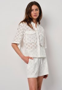 Witte oogjesstof outfit met een korte mouwen knoopshirt met twee borstzakken en bijpassende shorts; beiden hebben uitsnijdpatronen.