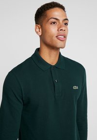 Temnozeleno dolgomajico s polo ovratnikom in gumbi, teksturiran material in majhna izvezena logotip na prsih.