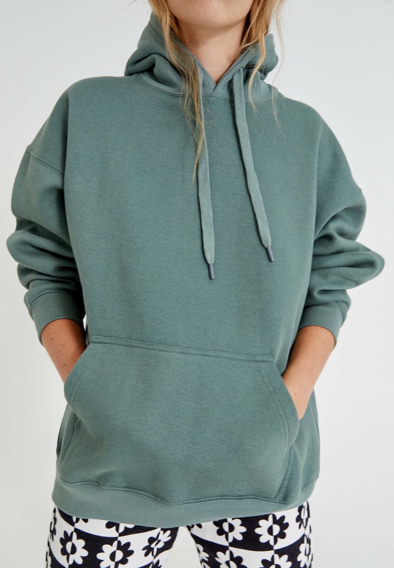 Personne portant un sweat à capuche vert avec des poches avant et un pantalon à motif floral noir et blanc sur un fond uni.