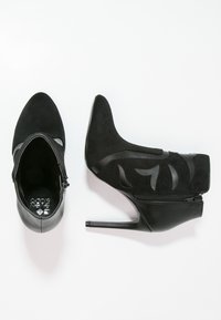 Botines negros de tacón alto con puntera puntiaguda, hechos de ante y cuero, con recortes decorativos y detalle de cremallera lateral.