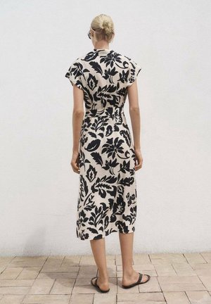 Femme aux cheveux blonds en chignon bas porte une robe midi beige à motif floral noir et des sandales slip-on noires, debout sur un sol carrelé.