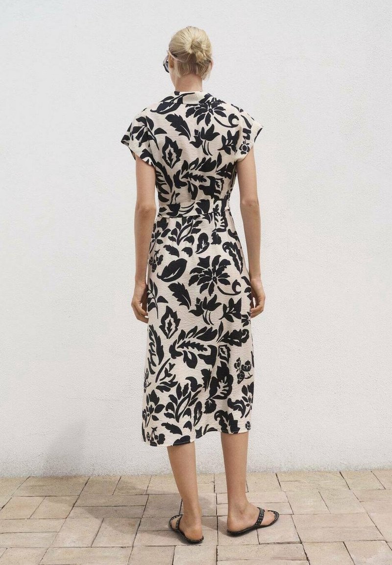 Femme aux cheveux blonds en chignon bas porte une robe midi beige à motif floral noir et des sandales slip-on noires, debout sur un sol carrelé.