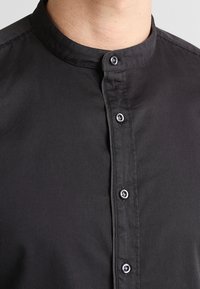 Camicia nera con bottoni, dotata di un colletto rotondo, texture liscia e cinque bottoni frontali, con una leggera lucentezza sul tessuto.
