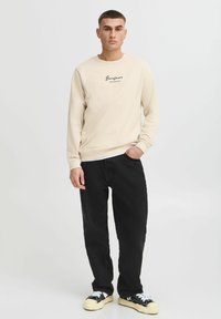 Felpa beige con logo nero a scritta, abbinata a pantaloni neri dalla vestibilità ampia e sneakers nere con suole bianche. Sfondo semplice.