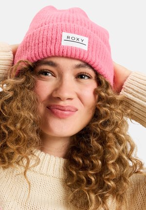 Berretto lavorato a maglia rosa con texture a coste e un'etichetta bianca con il marchio. Il modello indossa il cappello insieme a un maglione beige con lavorazione grossa.