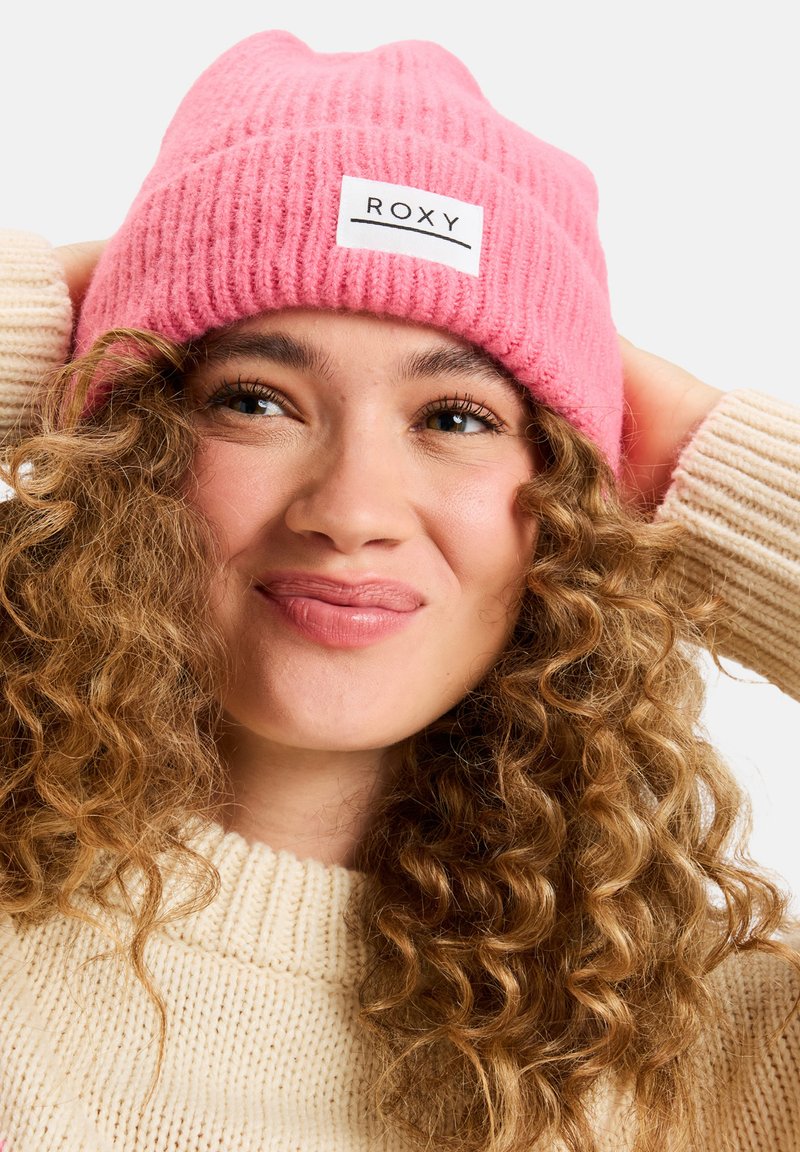 Różowy dzianinowy czapka z ribbowanym wzorem i białą metką z logo. Modelka prezentuje czapkę, nosząc sweter w kolorze kremowym z grubym splotem.