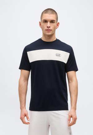 Mees, kes kannab tumesinist T-särki valge horisontaalse triibu ja EA7 logoga, kombineeritud valgete lühikestega, lihtsa taustaga.