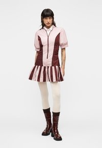 Φόρεμα με κοντά μανίκια και φερμουάρ στο μπροστινό μέρος, με ροζ τοπ και πτυχωτή φούστα με ρίγες σε μαρόν χρώμα. Το μοντέλο φοράει γυαλιστερά καφέ μποτάκια μέχρι το γόνατο.