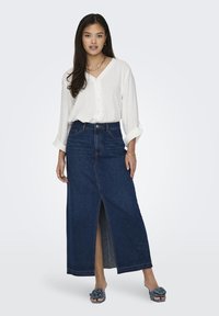 Cămașă albă cu buline, cu mâneci suflecate; fustă maxi din denim albastru închis, cu o deschidere frontală; sandale moi, gri, decorate cu flori.