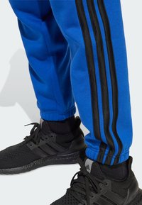 Un pantalon de survêtement bleu avec des bandes noires sur les côtés, associé à des baskets noires avec une tige en maille et une semelle texturée.