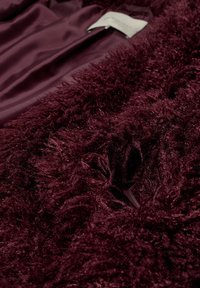 Burgundy fluffig tyg med en plyschig textur, med en slät satininsida och en liten etikett synlig i kanten.