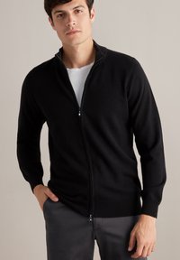 Svart zip-up cardigan i mjukt stickat material med ståkrage och långa ärmar, bärs över en vit t-shirt. Enkelt design, utan mönster.