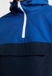 Blue and navy windbreaker with a zippered front, adjustable hood, and a chest pocket. Smooth texture with two-tone design.