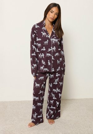 Femme debout pieds nus portant un pyjama violet foncé à manches longues avec des motifs de chien dalmatien blancs sur un fond clair.