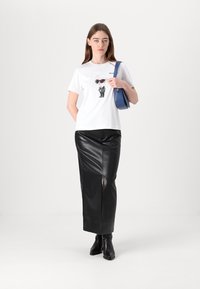 Weißes T-Shirt mit grafischem Design einer Figur in Sonnenbrillen, kombiniert mit einem figurbetonten schwarzen Leder-Maxirock und schwarzen Ankle Boots.