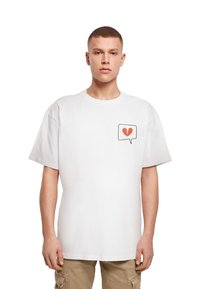 Bílá bavlněná tričko s krátkým rukávem, které má na levé straně hrudníku malou grafiku červeného zlomeného srdce uvnitř mluvící bubliny.