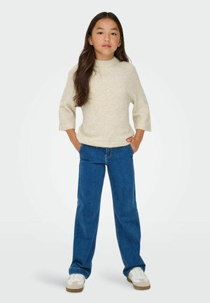 Lichtbeige, wollen trui met hoge nek en driekwart mouwen, gecombineerd met blauwe, rechte spijkerbroeken en witte sneakers.