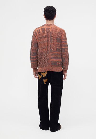 Homme de dos portant un pull texturé orange et marron à motifs et un pantalon noir avec des motifs d'oiseaux orange sur une poche arrière.