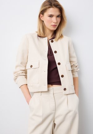 Joven mujer con chaqueta crema abotonada con bolsillos parche y pantalones a juego de talle alto, combinados con una blusa marrón oscuro.