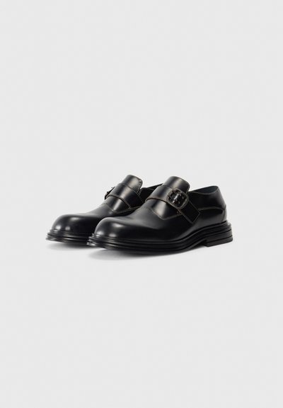 Une paire de chaussures en cuir verni noir avec boucles en argent et semelles superposées sur fond blanc.