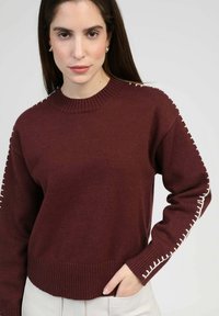 Pull en tricot couleur bordeaux avec un ourlet côtelé et des coutures décoratives blanches le long des manches. Col rond et coupe ample.