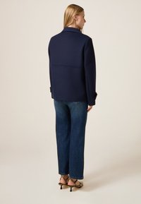 Mujer de espaldas vistiendo un blazer azul marino, jeans azules y sandalias de tacón con estampado de serpiente frente a un fondo beige liso.