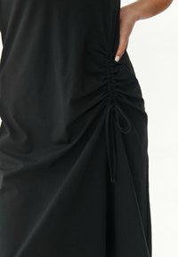 Schwarzes Stoffkleid mit geraffter Seite und verstellbaren Bindungen, weicher Textur, minimalistisches Design und fließender Silhouette.