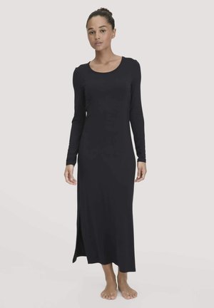 Donna in piedi a piedi nudi che indossa un vestito maxi nero a maniche lunghe con uno spacco laterale, su uno sfondo chiaro e semplice.