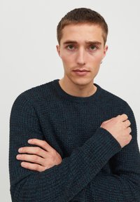 Mörkblå stickad tröja med ett strukturerat mönster, rund halsringning och ribbade ärmslut. Modellen står med armarna i kors och visar upp plaggets passform.