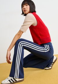 Piros kötött ujjatlan pulóver egy bézs testhezálló ing fölött, kék, bővülő nadrággal, fehér csíkokkal. A lábbeli virágos díszítéssel rendelkezik.