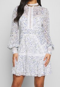 Žena v dlouhých rukávech, bílých květinových šatech s krajkovými detaily, sklady na těle a volánkovou sukní, stojící před neutrálním pozadím.