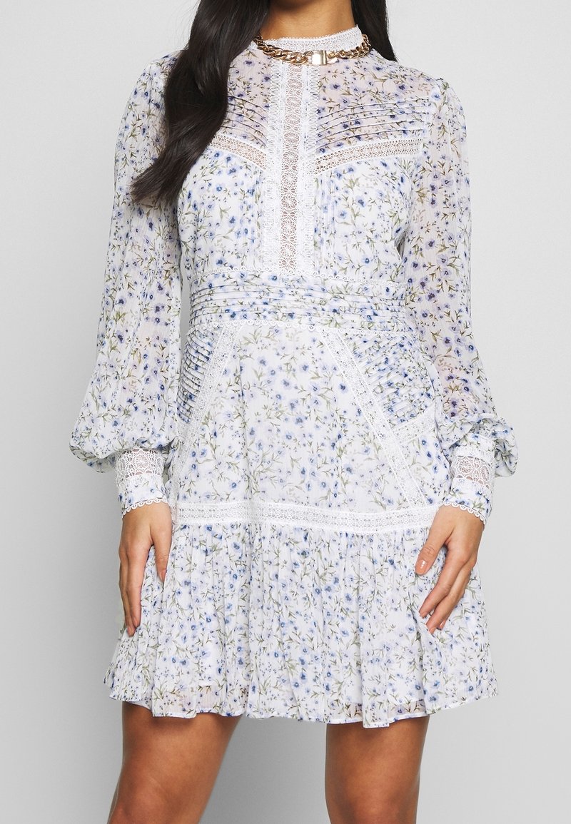 Žena v dlouhých rukávech, bílých květinových šatech s krajkovými detaily, sklady na těle a volánkovou sukní, stojící před neutrálním pozadím.