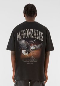 Sort oversized t-shirt med et fremtrædende �ørn grafik, lyn detaljer og teksten "M. GONZALES" i en slidt skrifttype på ryggen.