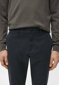 Mężczyzna ubrany w granatowe spodnie i brązowy sweter z długim rękawem, ręce swobodnie opuszczone wzdłuż ciała, z zapiętym paskiem widocznym w talii.