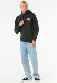 Felpa con cappuccio grigio scuro, dotata di una tasca frontale e di un logo circolare. Abbinata a jeans azzurri chiari e sneakers nere con suola piatta.