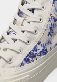 Λευκό ψηλό αθλητικό παπούτσι με μπλε λουλουδάτο σχέδιο, λευκά κορδόνια και χοντρή σόλα από καουτσούκ σε κοντινή λήψη.