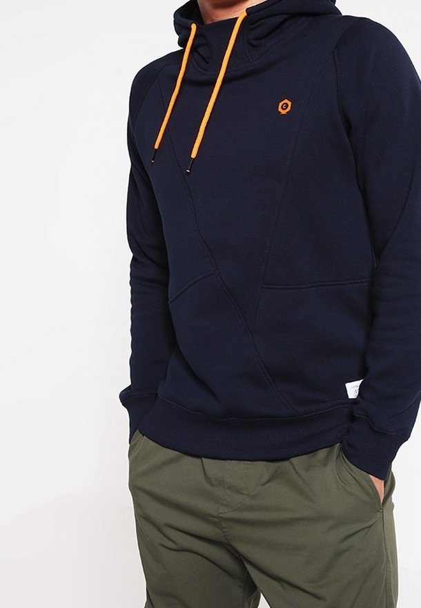 Personne portant un sweat à capuche bleu marine avec des cordons orange et un petit logo, et un pantalon vert olive, mains dans les poches sur un fond blanc uni.
