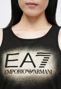 Fekete trikó, arany-fekete átmenetes logóval, rajta az "EA7 EMPORIO ARMANI" felirattal. Kerek nyakkivágás és ujjatlan kialakítás.
