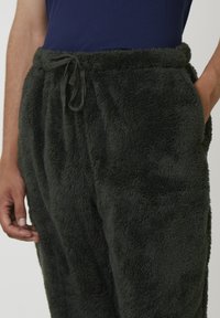Man som bär mörkgröna fluffiga fleecbyxor med dragsko i midjan och fickor, kombinerat med en marinblå texturerad skjorta.
