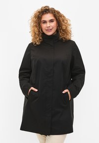 Black waterproof coat with high collar, two front pockets, and a straight cut. Smooth texture and minimalistic design with no visible hardware.