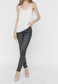 Kvinna i vit spetskantad linne, mörka skinny jeans och beige klackar, stående med ena handen i fickan mot vit bakgrund.