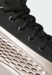 Černá sportovní bota s texturovaným textilním svrškem, plochými černými tkaničkami a kovově zlatou podrážkou s včelím plástem.