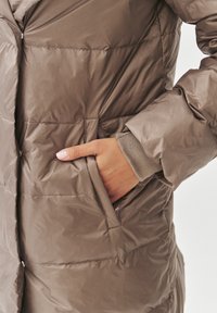 Bruine pufferjas met een zogenaamde quilted textuur, voorzien van een zijzak en gebreide manchetten. Gladde, glanzende stof met subtiele naden en accenten.